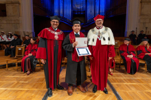 A male graduate in a cap and gown stands between two university officials in ceremonial robes and holds an open diploma folder in his hands while posing for a photograph.