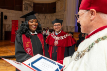A smiling woman in a graduation cap and gown receives a diploma folder from a university official. Another official in a ceremonial robe stands behind her.