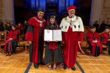 A woman in a graduation cap and gown poses between two university officials in ceremonial robes while holding an open diploma folder and a diploma certificate in her hands.