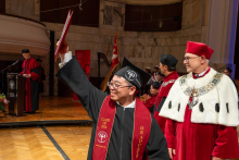 A male graduate in a cap and gown raises a diploma folder above his head as he celebrates receiving his diploma. University officials stand beside him.