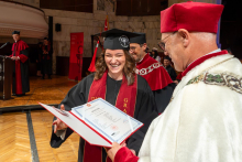 A smiling woman in a graduation cap and gown receives a diploma folder from a university official in a ceremonial robe.