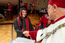 A woman in a graduation cap and gown shakes hands with a university official in a ceremonial robe. A diploma certificate is visible in the foreground.
