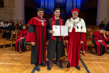 A male graduate in a cap and gown poses between two university officials in ceremonial robes while holding an open diploma folder and a diploma certificate in his hands.