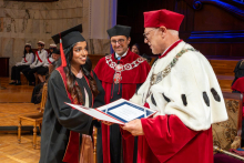 A woman in a graduation cap and gown receives a diploma folder from a university official. Another official in a ceremonial robe watches the ceremony.