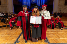 A smiling woman in a graduation cap and gown poses between two university officials in ceremonial robes while holding an open diploma folder and a diploma certificate in her hands.