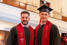 Two men in graduation gowns and red stoles stand side by side and pose for a photograph.