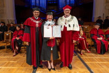 A smiling woman in a graduation cap and gown poses between two university officials in ceremonial robes while holding an open diploma folder and a diploma certificate in her hands.