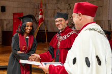 A woman in a graduation cap and gown shakes hands with a university official in a ceremonial robe while receiving her diploma.