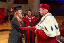 A woman in a graduation cap and gown shakes hands with a university official in a ceremonial robe while holding a diploma folder.