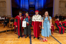 A male graduate in a cap and gown stands with two university officials in ceremonial robes and a woman in a blue dress. He holds a diploma and a gift bag in his hands.