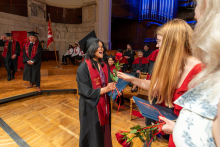 A woman in a graduation cap and gown receives a red rose from another woman. She holds a diploma folder in one hand and looks surprised and happy.