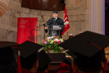 A man in an academic gown stands at a lectern and delivers a speech. A large floral arrangement and the faculty banner are placed nearby.