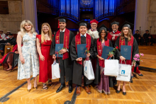 Three graduates in caps and gowns pose with two women and two university officials. The graduates hold diploma covers, flowers and gift bags in their hands.