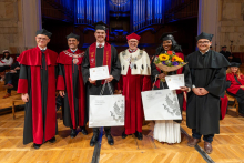 Two graduates stand with university officials and academic staff. The man holds a certificate and a gift bag, while the woman holds a bouquet of flowers, a certificate and a gift bag.