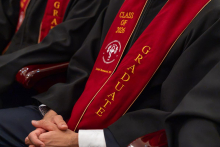 A close-up of a graduate’s hands resting on their lap. A red stole embroidered with the words “Class of 2026” and “Graduate” is draped over the academic gown.