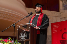 A male graduate in a cap and gown stands at a lectern and delivers a speech during the graduation ceremony.