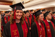 A smiling woman in a graduation gown and cap stands among a group of graduates seated in an auditorium.