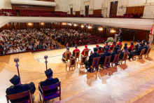 A wide view of the graduation ceremony in a concert hall. Graduates and university officials sit on stage while a speaker addresses a large audience.