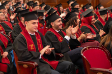 Male and female graduates in academic gowns, caps and red stoles sit in an auditorium and applaud during the graduation ceremony.
