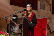 A woman in a graduation gown, cap and glasses stands at a lectern and delivers a speech. A faculty banner is displayed behind her.