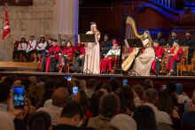 Two female musicians perform on stage. One woman plays the violin and the other plays the harp while university officials and graduates sit behind them.