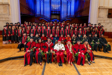A large group of graduates and university officials pose for a commemorative photograph on stage. Many of them hold white roses in their hands.