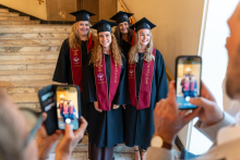 Four women in graduation gowns and caps pose for a photograph near a marble staircase. Two people hold mobile phones in their hands while taking their picture.