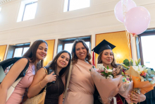 Three women pose with a female graduate in a graduation cap and gown. The graduate holds bouquets of flowers in her arms, and pink balloons float above the group.