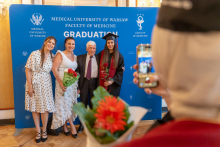 A female graduate in a graduation gown and cap poses with three family members in front of a university backdrop. She holds a red rose in her hand, while two women hold bouquets of flowers.