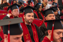 A man in a graduation gown and cap looks toward the camera while seated among other graduates wearing academic attire.