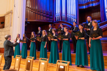 A university choir performs on stage. Women and men dressed in formal black and green concert attire hold music folders in their hands while a man conducts the ensemble.