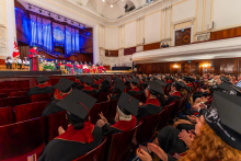 Graduates in academic gowns sit in an auditorium facing a stage where university authorities are seated and a speech is being delivered.