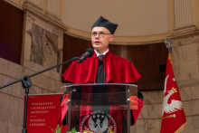 A man in a red academic robe speaks at a lectern equipped with a microphone. A university banner and the Polish flag stand behind him.