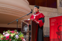 A man in a ceremonial academic robe stands at a lectern and delivers a speech. A large floral arrangement is placed in front of the stage.
