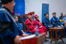 Several people in academic regalia are sitting on stage during the ceremony. In the foreground, a person is seen speaking at the podium.