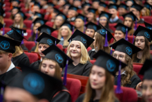 Uczestnicy uroczystości akademickiej siedzą w auli w togach i biretach. W kadrze widoczne są kolejne rzędy osób biorących udział w wydarzeniu.