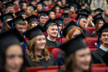 The participants in the academic ceremony are seated in the auditorium wearing academic gowns and caps. The frame shows a large crowd gathered in the audience.