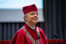Mężczyzna w stroju akademickim stoi przy mównicy i przemawia podczas uroczystości. W tle widoczny jest ekran oraz elementy wyposażenia sceny.