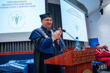 A man in academic regalia is standing at the podium, clapping his hands. In the background, a screen displays the name of the event, the people seated on stage, and the diplomas that have been prepared.