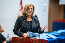 A woman is standing at the podium, giving a speech. She is holding some documents in her hand. In the background, you can see the diplomas that have been laid out.