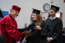 A woman receives her diploma during an academic ceremony. In the background, other participants can be seen waiting to receive their diplomas.