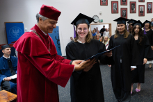 During the diploma ceremony, a person in academic regalia presents the document to a participant in the ceremony. The next people are waiting in line.