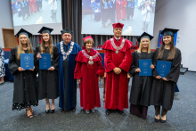 Four people in academic gowns and caps are holding diplomas and posing for a group photo with three people in academic regalia. In the background, projection screens display footage from the ceremony.