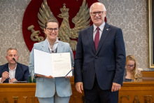 Dwie osoby pozują do zdjęcia. Jedna z nich trzyma otwartą teczkę z dokumentem. W tle widoczne są osoby siedzące przy stole prezydialnym oraz godło.