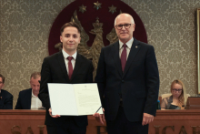 Dwie osoby stoją przed godłem. Jedna z nich prezentuje otwarty dokument. W tle znajdują się uczestnicy obrad siedzący przy stole.