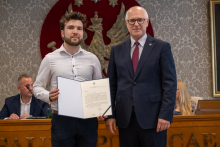 Dwie osoby stoją obok siebie. Jedna trzyma otwartą teczkę z dokumentem skierowanym do aparatu. W tle widoczne są osoby uczestniczące w posiedzeniu.