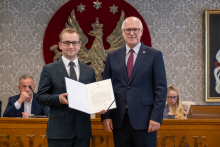 Dwie osoby pozują do zdjęcia z otwartym dokumentem. Za nimi znajdują się uczestnicy spotkania oraz stół prezydialny.