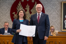 Dwie osoby stoją przed godłem. Jedna z nich prezentuje otwartą teczkę z dokumentem. W tle widoczne są osoby siedzące przy stole.