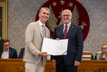 Dwie osoby stoją obok siebie i trzymają otwarty dokument. W tle znajdują się uczestnicy posiedzenia oraz elementy wyposażenia sali obrad.