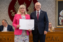 Dwie osoby pozują do zdjęcia. Jedna prezentuje otwartą teczkę z dokumentem. W tle widoczne są osoby uczestniczące w spotkaniu.